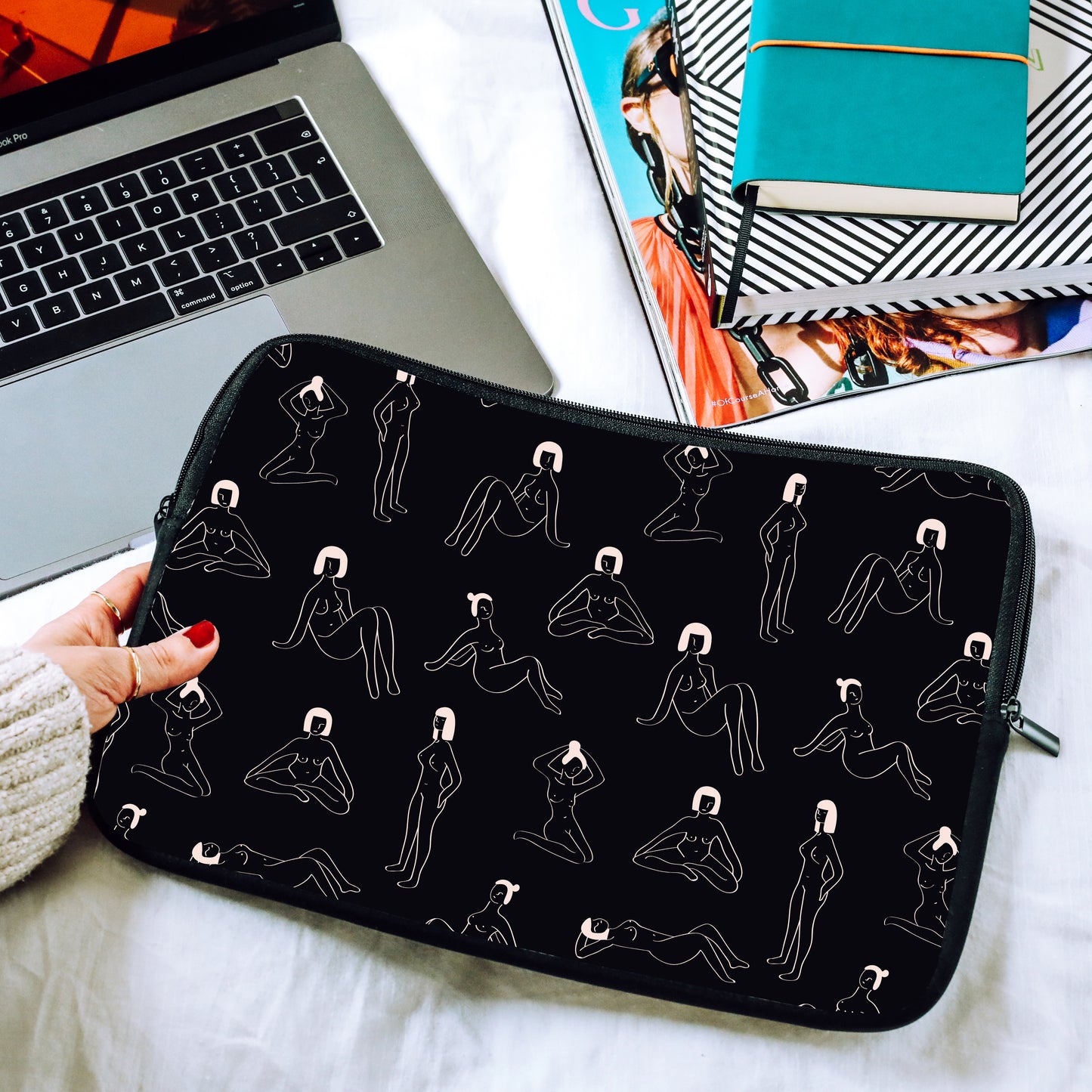 Hand holding a black laptop sleeve with naked ladies design on a bed with a laptop and books in the background | Harper & Blake