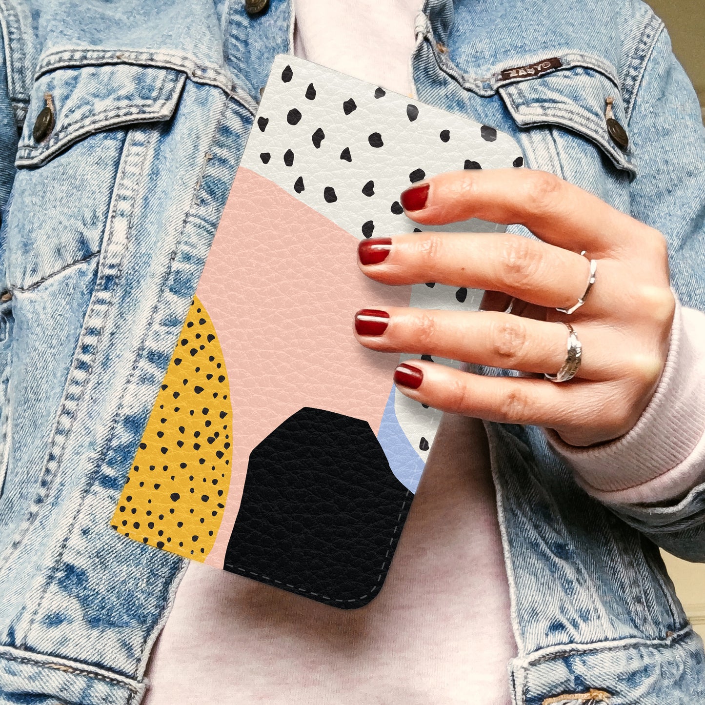 A woman holding a wallet phone case and wearing a light denim jacket. The wallet case has polka dots and pink, black and yellow shapes. 