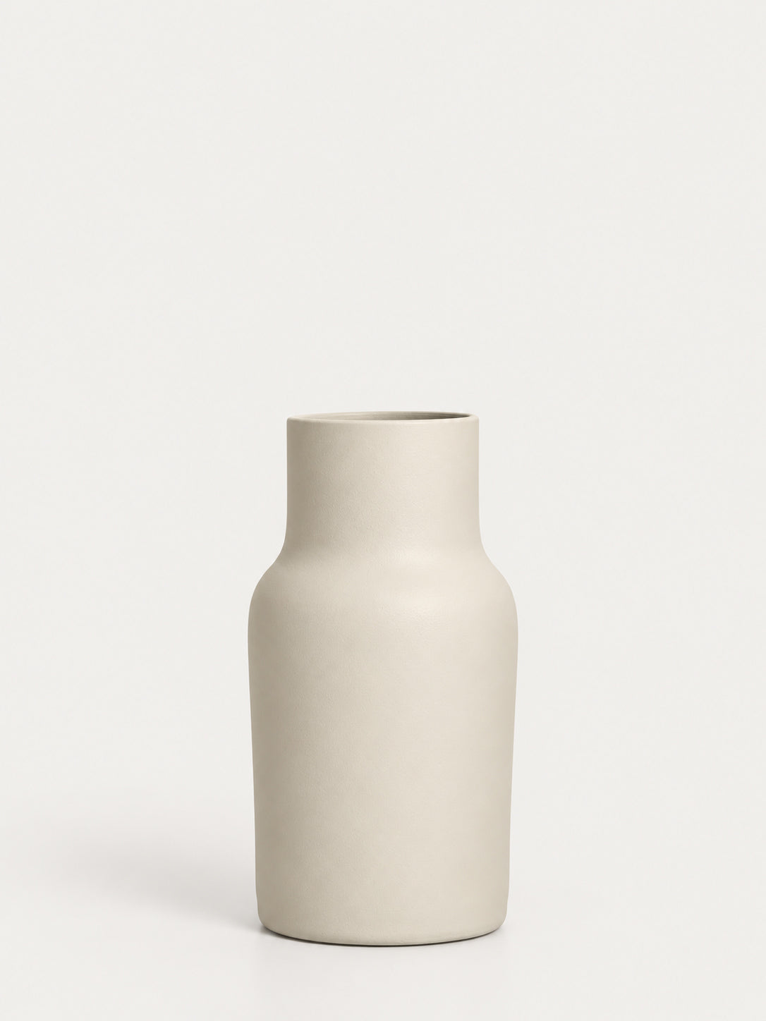 A minimalist ceramic vase with a matte, light beige finish, standing on a clean white surface against a solid off-white background.