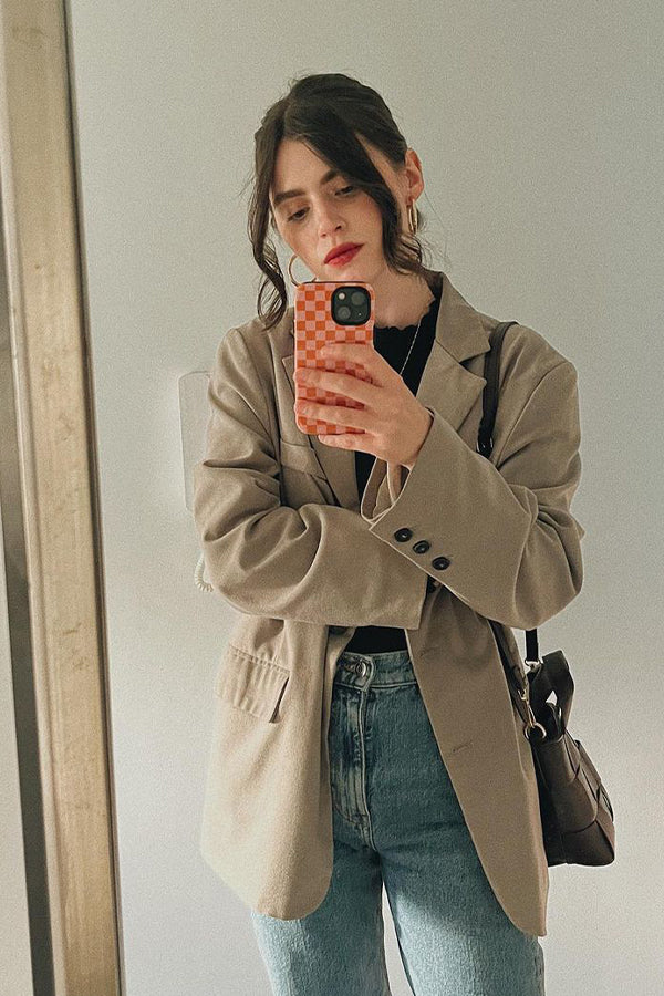 Girl doing a mirror selfie wearing a brown jacket and jeans. The phone case is pink and orange in a check pattern