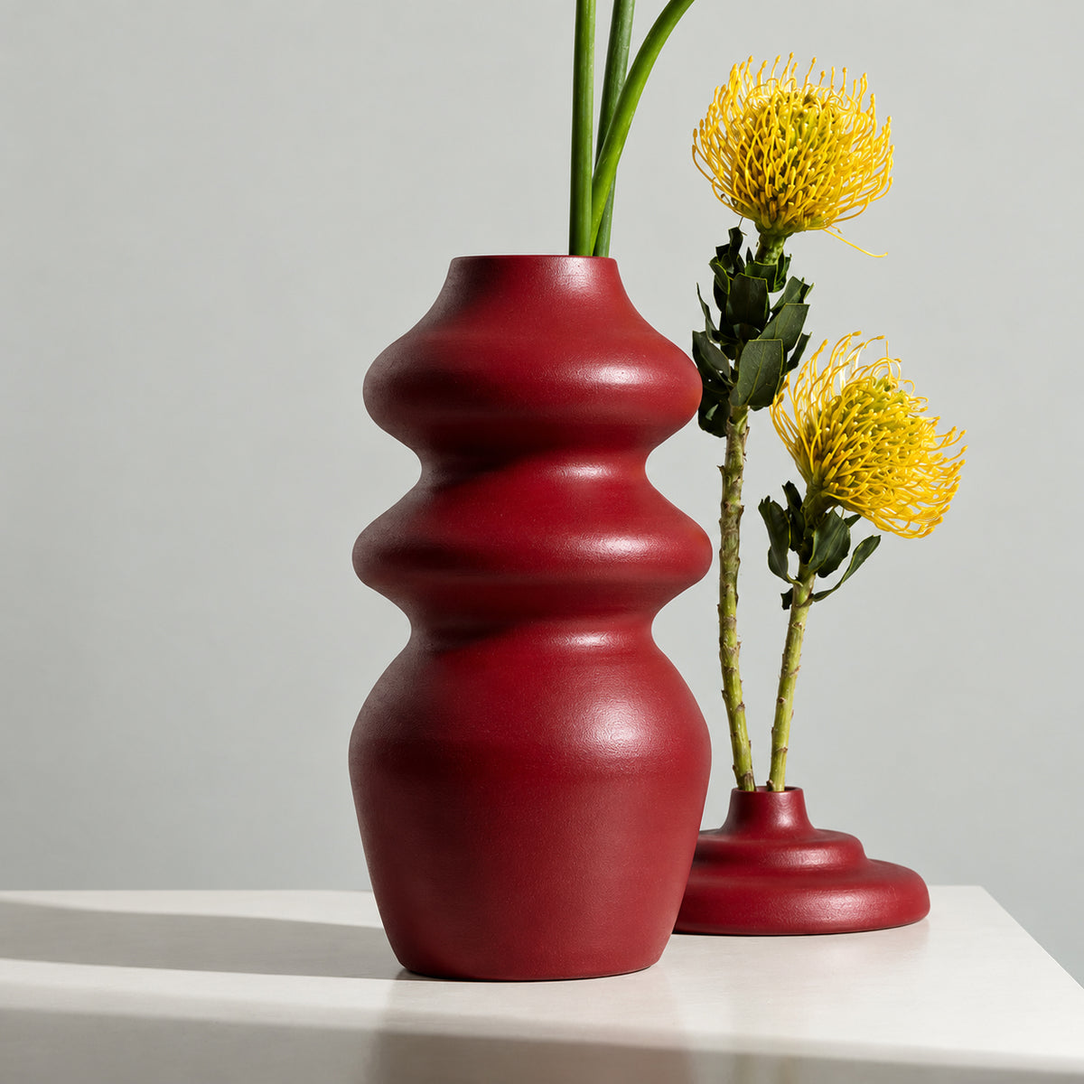 A glossy deep red ceramic vase with a rounded, segmented organic shape stands on a white surface against a light gray background. A second matching red vase holds two bright yellow flowers.