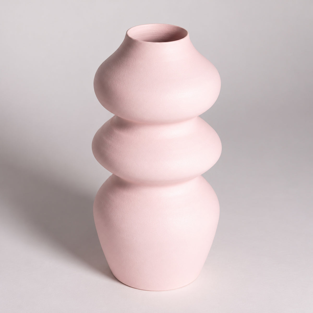 A light pink ceramic vase with a stacked, rounded three-tiered design, photographed on a neutral light gray background.