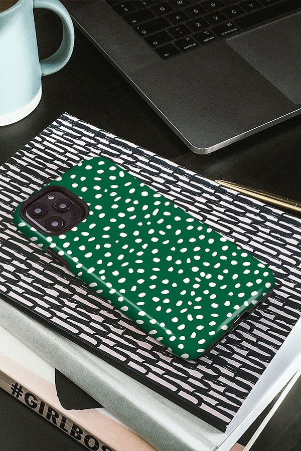 Green phone case with white polka dots on a black and white patterned surface, with a laptop and mug in the background.