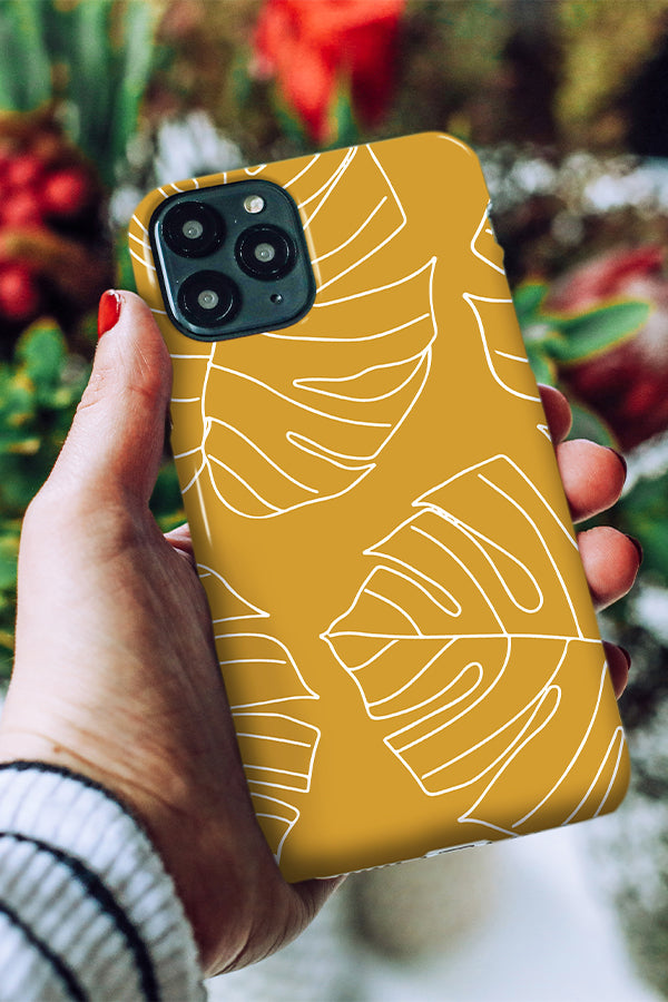 Hand holding a yellow phone case with white leaf patterns against a blurred natural background