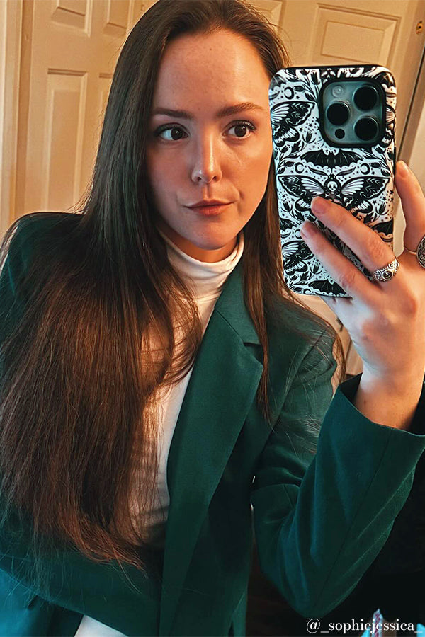 Woman holding a phone taking a selfie. She's wearing a green blazer and the phone case has a moth design on it 