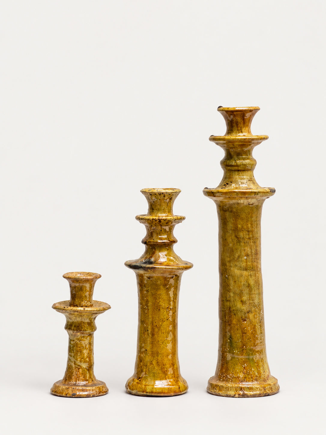 Three ceramic candlesticks in graduated sizes, featuring a glossy ochre and brown glaze with subtle greenish-yellow undertones, set against a plain white background.