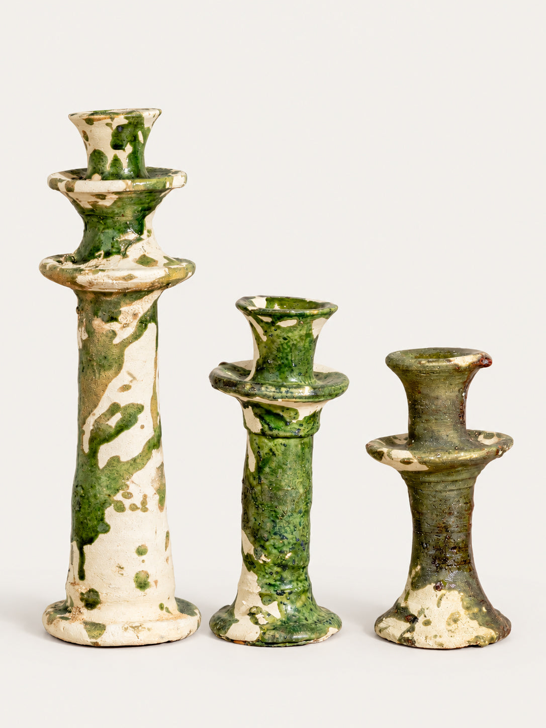 Three ceramic candlesticks of varying heights with a distressed green and cream glaze, arranged from tallest to shortest against a plain off-white background.
