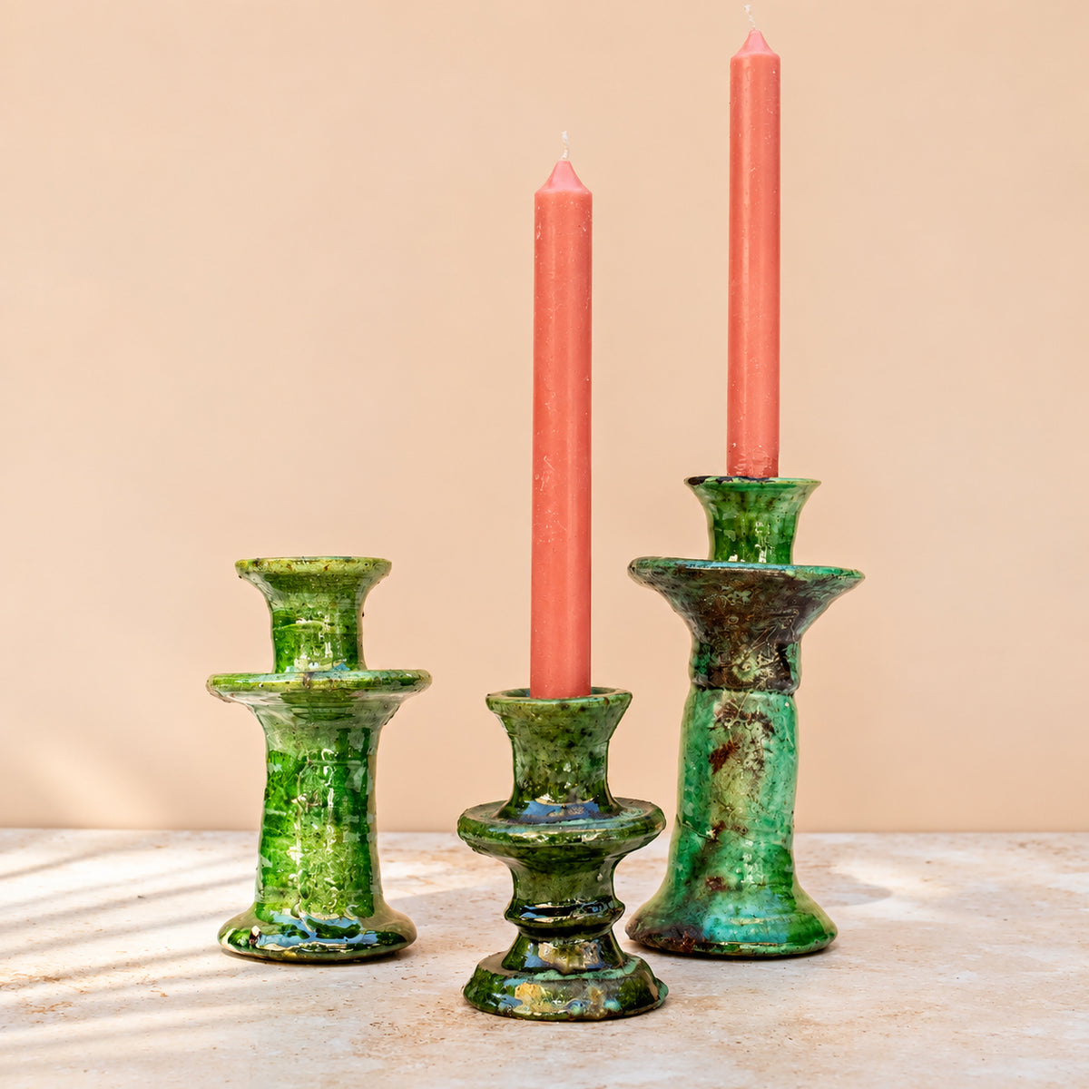 Three green glazed ceramic candlesticks on a light stone surface against a warm beige background, with two coral-pink taper candles inserted.