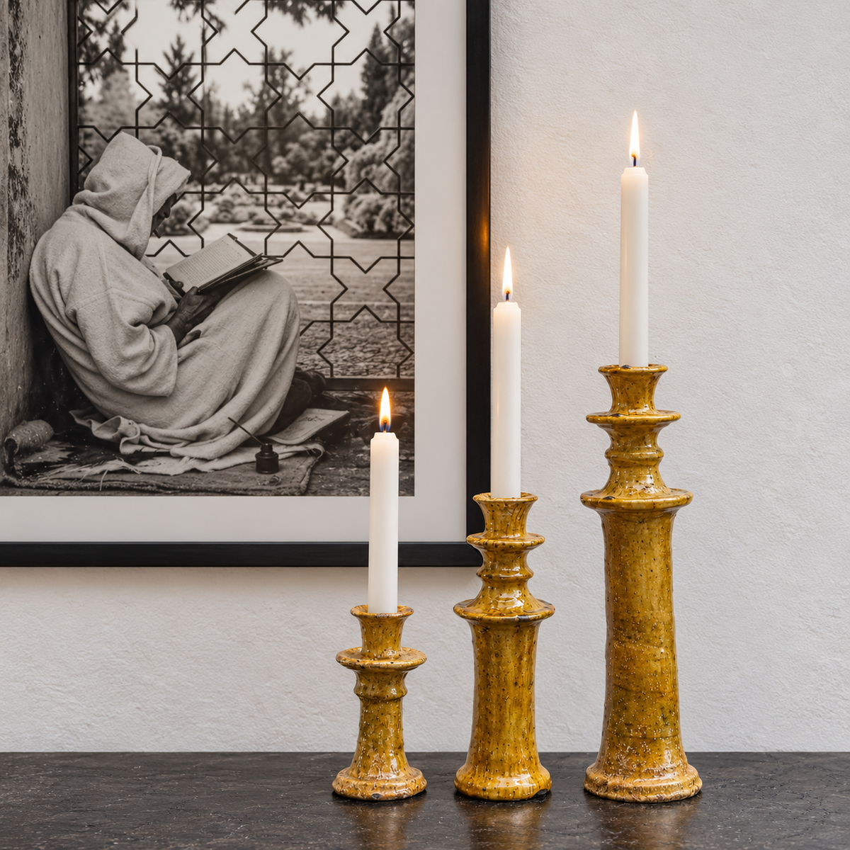 Three gold ceramic candlestick holders of varying heights holding white taper candles, styled on a dark tabletop in front of a framed black-and-white photograph against a textured off-white wall.