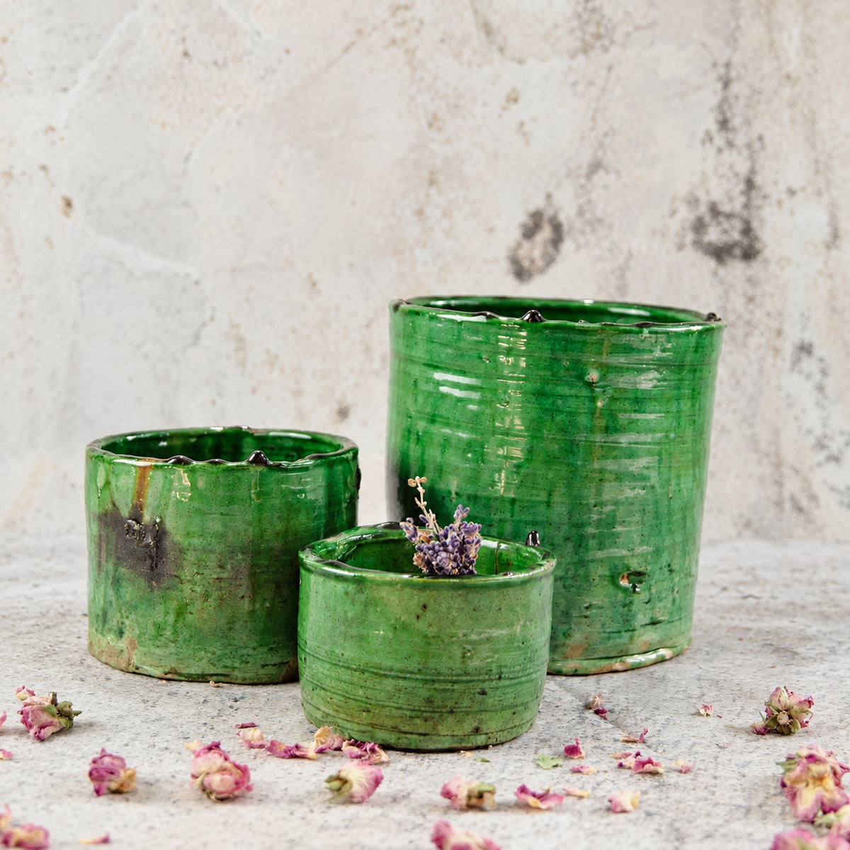 Three handmade green glazed ceramic planters of varying sizes, arranged on a textured light gray surface with scattered dried pink flowers. The largest planter is in the back, followed by a medium planter, and a small planter in the front containing a small sprig of dried lavender.