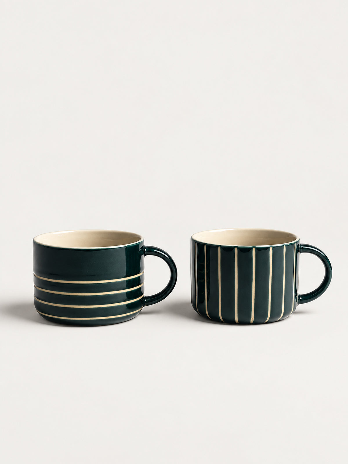 Two dark green ceramic mugs with cream interiors, one featuring horizontal cream stripes and the other vertical cream stripes, shown on a light gray background.