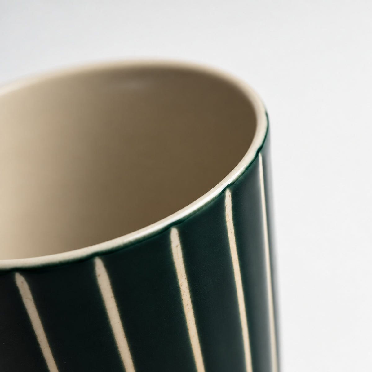 Close-up of a ceramic mug showing its dark green exterior with vertical cream stripes and the beige interior, set against a plain light gray background.