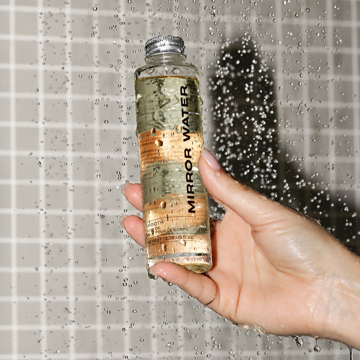 A hand holds a clear glass bottle labeled 'MIRROR WATER' filled with a golden liquid, covered in water droplets, against a tiled wall with water spraying behind it.