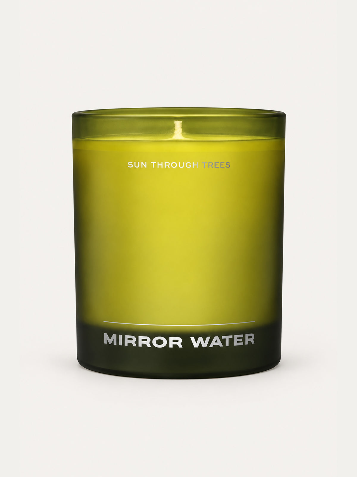 A candle in a translucent olive-green glass jar with a black base, featuring white text that reads 'SUN THROUGH TREES' and 'MIRROR WATER'.