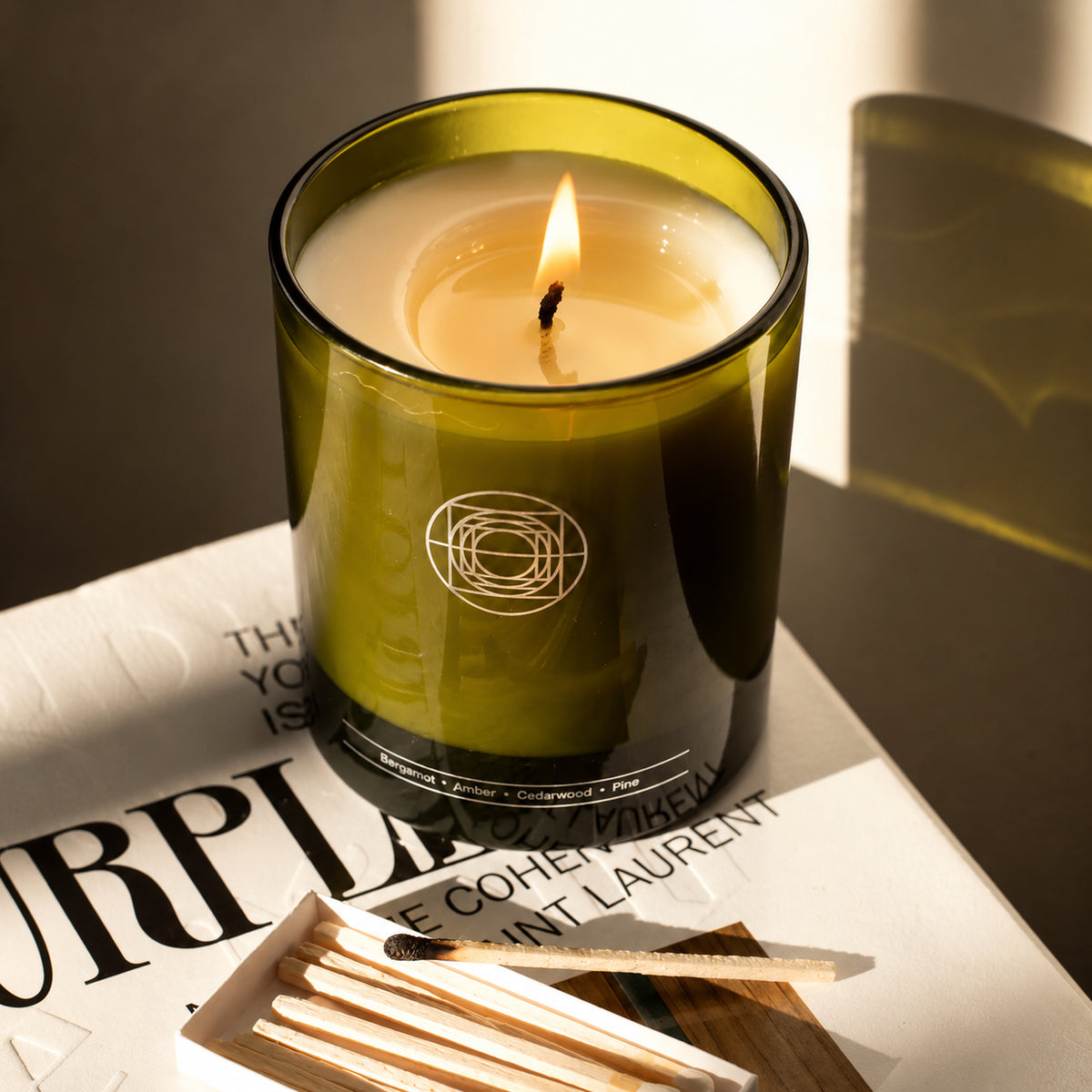 A lit candle in an olive green glass jar sits on a white book with printed text, alongside a small matchbox. The candle has cream-colored wax and a visible flame, with warm sunlight casting shadows across the scene.