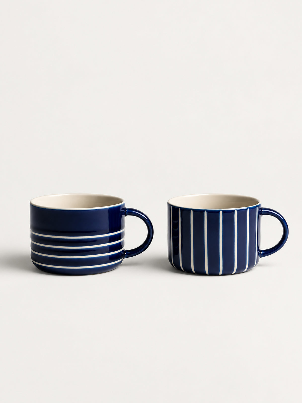 Two navy blue ceramic mugs with white stripes, one featuring horizontal stripes and the other vertical stripes, set against a plain light background.
