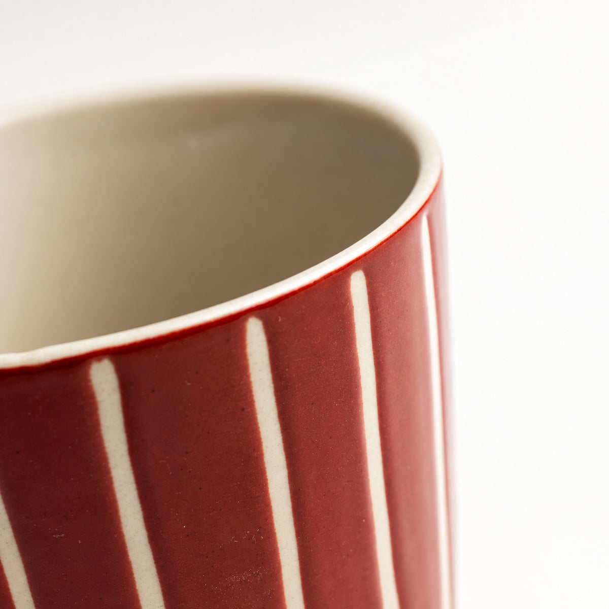Close-up of a ceramic mug showing a deep red exterior with vertical white stripes and a light beige interior, set against a plain white background.