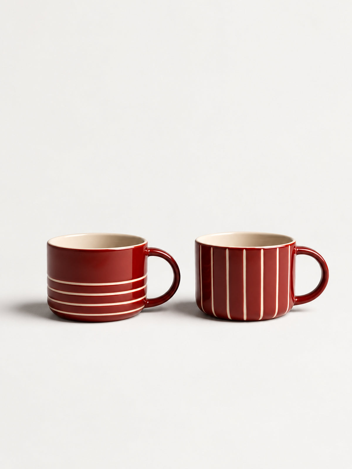 Two red ceramic mugs with cream-colored stripes, one featuring horizontal stripes and the other vertical stripes, set against a plain light gray background.